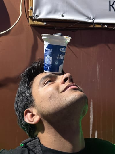 The image shows a close-up, low-angle shot of a young man balancing a plastic cup on his forehead. The man has dark, curly hair and is looking upwards with his mouth slightly open. The cup, labeled in Cyrillic with the word "Айрян" (Ayran), is white with blue lettering and features an image of milk. The background is a textured, reddish-brown wall with a white banner stretched across the top, attached with black zip ties and metal grommets. A white electrical cord runs diagonally across the upper left of the frame. The scene appears to be outdoors, possibly in a city like Razgrad, Bulgaria, given the Cyrillic labeling on the cup. The lighting suggests it is either evening or a shaded area, with a faint green glow on the man's neck and shoulder.