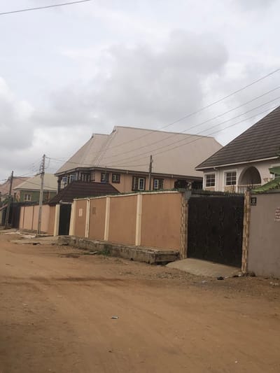 The scene depicts a residential street in Egan, Nigeria, under an overcast sky. The foreground features an unpaved, reddish-brown dirt road with visible tire tracks and scattered debris. A damaged concrete drainage channel runs along the right edge of the road. Further right, a dark, ornate metal gate, flanked by columns with stone tile accents, is integrated into a light-colored perimeter wall. A house number "18" and a partially obscured sign are visible on this wall. Behind the wall, a two-story building with a light beige exterior, multiple windows, and a large tan corrugated metal roof with prominent gables is situated. To its immediate right, another house with a dark brown tiled roof and a white facade featuring an arched entrance is visible. On the left side of the road, a long, peach-colored perimeter wall with vertical white columns extends into the background. Behind this wall, a building with a dark brown roof is partially visible. Further down the street, another residential structure is faintly discernible. Overhead utility poles with multiple power lines traverse the scene. No individuals, actions, or interactions are present.