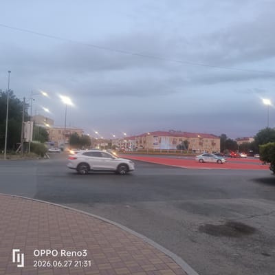 The image depicts a street scene in Atyrau, Kazakhstan, under a twilight sky with scattered clouds. The foreground features a paved sidewalk and a road. Several vehicles are visible, including a white SUV in motion on the road, a police car, and other cars in the distance. Buildings line the street, with some displaying signs. A fountain is also visible in front of one of the buildings. Streetlights illuminate the area. The timestamp "2026.06.27 21:31" and "OPPO Reno3" are overlaid on the bottom left corner of the image.
