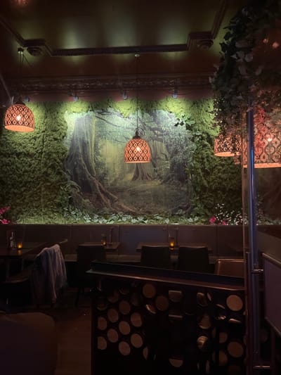 The interior of a restaurant located at coordinates 48.201, 16.362 in Vienna, Austria, featuring a large mural depicting a lush forest scene. The mural is partially covered with moss and greenery. The restaurant has a dark wood aesthetic with several booths lined against the wall. Each booth has a dark wood table and chairs. There are several hanging light fixtures with ornate designs. The lighting is dim, creating a cozy and intimate ambiance.  The scene is viewed through a partially obscured doorway, with a dark, patterned screen partially visible in the foreground.  The only discernible person in the scene is a figure sitting in a booth, with a denim jacket hung on the back of the chair.  The restaurant's ambience is relaxed and inviting.