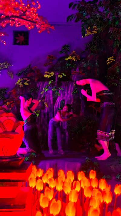 Captured in Binh Lu, Vietnam, the image reveals a scene with three figures set against a backdrop mimicking a waterfall surrounded by trees and foliage. The artificial waterfall, illuminated in shades of purple, cascades down a wall. To the left, a female mannequin stands with her hands behind her head, partially obscured by a large red floral decoration in the foreground. Next to her is a female individual, dressed in a light-colored top and jeans, leaning towards the waterfall. On the right stands another, taller female mannequin, adorned in traditional attire, her hands positioned near her face. The foreground is dominated by a cluster of glowing orange tulip-shaped lights. The scene is under artificial lighting, enhancing the colors and creating a theatrical atmosphere. In the upper left corner, a square vent or window is visible, adding to the artificiality of the setting.