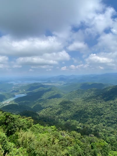 The image shows a panoramic view of a mountainous, forested landscape with bodies of water, likely reservoirs or lakes, visible in the distance. The foreground is dominated by lush green trees and vegetation. The sky is partly cloudy, with a mix of blue sky and white clouds. The scene is characterized by rolling hills covered in dense forest, with some of the hills appearing as dark shadows due to the lighting. In the mid-ground and distance, the terrain becomes softer and more undulating, with layers of mountains fading into a hazy blue horizon. A few roads or pathways might be discernible within the treeline in some areas. The overall impression is one of a serene and natural environment. The prompt specifies the location as Phan Son, Vietnam, which implies this is a scenic viewpoint overlooking the region. There are no people or specific objects of interest other than the natural landscape.