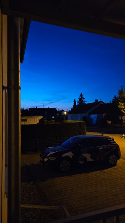 The image shows a street scene at dusk in Vöhringen, Germany. A dark-colored car with a camouflage-like design on its side is parked on a paved driveway. In the background, there are several houses with visible roofs and windows. A tall, dark green hedge runs across the middle ground of the image, separating the driveway from the houses. The sky is a deep blue, indicating either early morning or late evening. A downspout is visible on the left side of the frame, attached to a building that is partially illuminated by yellow light.