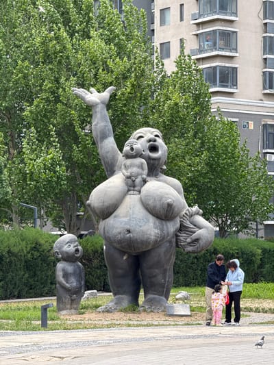 The image captures an outdoor scene in Changping District, China, featuring a large, grayish-bronze sculpture of a woman with multiple babies attached to her body. The sculpture, characterized by exaggerated features and open-mouthed expressions, stands prominently amidst greenery. In the background, a multi-story building with windows is visible, and a few individuals, including a woman, a man, and a young child in pink pajamas, stand on a paved walkway near the sculpture. A single bird is perched on the edge of the walkway. The environment includes trees, bushes, grass, and a patterned sidewalk.