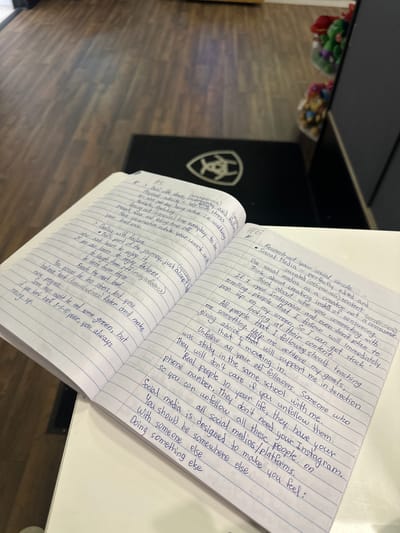 The image shows a close-up of an open notebook with handwritten notes, lying on a white surface. The notebook is filled with blue ink and is lined. The left page has handwritten lists and bullet points, some marked with asterisks and numbers. The right page contains more extensive handwritten notes in a similar style. The notebook appears to be placed on a desk or table.
In the background, out of focus, a dark surface can be seen, possibly a desk or a shelf, with some colorful objects in clear plastic bags. These objects appear to be small toys or candies. A part of a wooden floor is visible in the lower left corner of the image.
The handwriting in the notebook discusses topics such as dealing with stress, physical activity, meditation, dealing with failure, and social media. There are also some English and Russian words present in the text, suggesting the writer might be multilingual. The content seems to be a personal reflection or a set of personal goals. The overall scene is casual and domestic, likely an indoor setting, possibly a home or study area. There are no people visible in the image. The lighting suggests it could be daytime, but the specific time or weather cannot be determined.