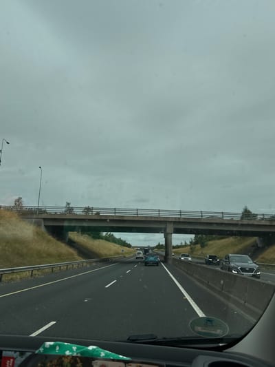 The image is taken from inside a vehicle, looking forward on a multi-lane highway under a cloudy sky. A concrete overpass stretches across the highway. On the overpass, there are railings and some vegetation on top. On the sides of the highway, there are grassy embankments and guardrails. Several cars are visible on the highway, traveling in the same direction as the camera's vehicle. One car, a dark SUV, is in the right lane. Another car, a light-colored sedan, is in the center lane, and a blue car is in the left lane. The scene appears to be within the vicinity of Moone, Ireland, as indicated by context. The road surface is asphalt, with white lane markings. The overall environment is that of a typical highway setting.