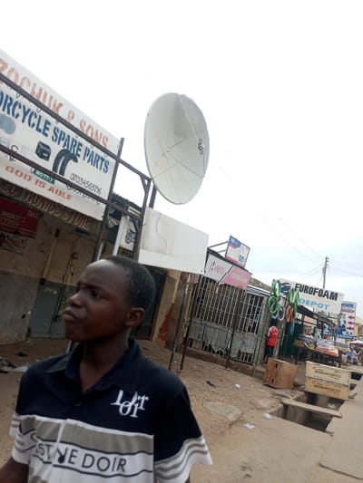 A low-angle medium shot shows a young boy looking to his left in Arawa, Nigeria. The boy is wearing a black polo shirt with white and gray stripes across the chest and a white collar. The background is a bustling street scene with various shops and businesses. One shop sign reads "IZOCHUK & SONS MOTORCYCLE SPARE PARTS". Another sign is for "EUROFOAM DEPOT". A satellite dish is mounted on a pole above the shops. In the foreground, there are some discarded papers on the ground. The street appears to be unpaved and dusty. The overall impression is one of a busy commercial area in a developing city.