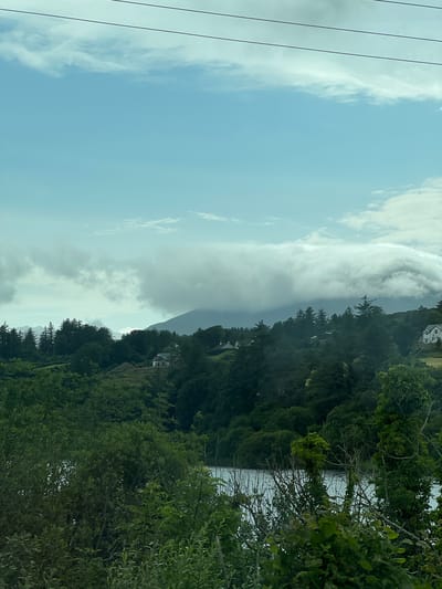 The image shows a scenic view of a cloudy sky over a body of water, with lush green trees and vegetation lining the shore. The perspective is from a high vantage point, looking down towards the water and the wooded hillside. There are a few houses visible nestled amongst the trees on the slope. The sky is partly cloudy with dramatic formations of white and grey clouds, suggesting a dynamic weather pattern. The time of day appears to be daytime, possibly late afternoon or early evening, given the soft lighting. The location is identified as Creeslough, Ireland, which is consistent with the verdant, coastal landscape. The overall mood is serene and natural, highlighting the beauty of the Irish countryside. There are no visible people or objects that suggest specific activity, but the presence of houses indicates a residential area within this natural setting. The image evokes a sense of tranquility and the raw beauty of nature.