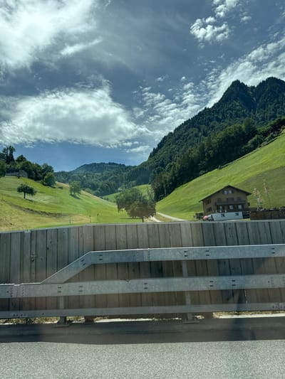 This image depicts a scenic view of a mountainous landscape in Beckenried, Switzerland. In the foreground, a grey asphalt road is visible, bordered by a wooden and metal guardrail. Beyond the guardrail, a lush green hillside slopes upwards, dotted with trees. A rustic, multi-story house with a brown facade and white window frames sits nestled on the hillside. Further in the background, dense green forests cover the steep mountain slopes, reaching towards a sky filled with dramatic, white cumulus clouds against a backdrop of blue. The overall impression is one of a tranquil, rural setting in the Swiss Alps.