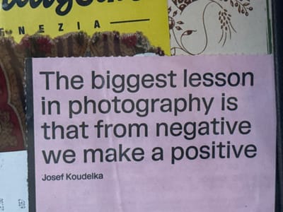 The image presents a close-up view of a static arrangement of layered papers and materials affixed to a surface in Cortona, Italy. Dominating the center is a light pink rectangular sheet of paper, approximately two-thirds the height of the frame, featuring black sans-serif text. The text reads: "The biggest lesson in photography is that from negative we make a positive," attributed to "Josef Koudelka" at the bottom. The top edge of the pink paper exhibits a slightly uneven, torn appearance.

Above the pink paper, a dark brown, textured strip of material with a roughly torn upper edge runs horizontally. Above this strip, to the upper left, a fragment of yellow paper displays the partial black text "VENEZIA." To the upper right, an off-white material or paper with a brown, intricate, stylized organic design, resembling a profile or floral pattern, is visible.

To the immediate left of the pink paper, a dark, patterned surface with hints of red and beige is partially visible, appearing to be fabric or decorative paper. Below this, a small white slip of paper with partial black numbers "12,00" and "6957" is exposed. The composition suggests a public display or a collection of layered, possibly weathered, documents or posters. No individuals, actions, or interactions are depicted.