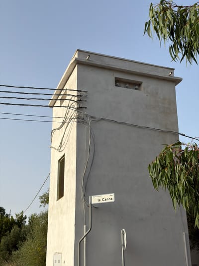 The image shows a light gray, rectangular, multi-story structure, possibly a utility building or watchtower, in Santu Diadòru/San Teodoro, Italy. The building has a flat roof with a short parapet and a small pipe protruding from the top. A rectangular window with a metal grate is visible on the left side of the building. Several electrical wires run horizontally across the upper left portion of the image and are attached to the building with various brackets and conduits.
On the front of the building, a white street sign is mounted on a gray pole. The sign reads "Burgeta la Suaredda Via la Canna". Below the window, a white electrical box with a grated cover is mounted on the wall.
The building is situated outdoors, with a clear blue sky in the background. Lush green foliage from a tree with long, narrow leaves hangs into the frame from the upper right and lower right portions of the image. The foliage appears to be eucalyptus. In the distance, to the left, there are more trees and vegetation, suggesting a rural or semi-rural setting within Santu Diadòru/San Teodoro. The lighting suggests it is daytime, possibly late afternoon, given the shadows. No people are visible in the image.
