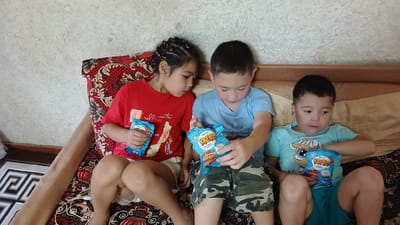 Three children are sitting on a couch in an indoor setting. The child on the left, a girl with braided hair, wears a red t-shirt and white shorts. She holds a blue snack bag. Beside her, a boy in a blue t-shirt and camouflage shorts holds another blue snack bag, looking down at it. On the right, a boy in a light blue t-shirt with a cartoon character and blue shorts also holds a blue snack bag. The couch has a patterned red and black fabric. The background consists of a textured wall and a wooden headboard. The scene appears to be in a domestic environment, possibly in Fergana City, Uzbekistan, given the cultural context.