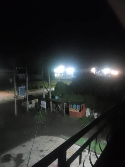 The scene captures a nighttime view in Njoro ward, Kenya. The perspective is elevated, with a dark metal balcony railing present in the bottom right foreground, overlooking a concrete surface. An unpaved road occupies the left midground, bordered by a utility pole assembly featuring a large electrical transformer and associated wiring. To the right of the road, a collection of makeshift commercial structures, including a reddish-brown kiosk with light-colored panels and other stalls partially obscured by vegetation and stacked items, are visible. A white rectangular sign is affixed to a pole near these structures. The background is characterized by multiple clusters of intense artificial illumination, indicative of distant buildings or establishments. The overall environment is dark, consistent with nighttime, with no discernible human presence or activity.