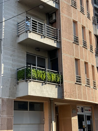 A low-angle, eye-level exterior shot captures a segment of a multi-story building in Vracar Urban Municipality, Serbia. The building's facade exhibits a mix of textures and colors. The left side of the building features a light-colored, textured concrete wall with horizontal grooving. On this section, a balcony with a metal railing and a trough of lush green plants is visible. Above this balcony, another balcony with a similar metal railing is seen. The right side of the building is characterized by rectangular panels of a reddish-brown, textured material separated by white trim. This section has multiple windows with metal shutters.
Below the balconies and windows, the ground level of the building reveals a business establishment with a glass storefront. Signage on the storefront includes "Intesa" and various icons. The entrance appears to be a commercial door. A metal garage door is also visible on the far left side of the image, at the base of the building. The lighting suggests it is daytime, with shadows cast across the building's facade. The overall scene depicts an urban architectural environment.
