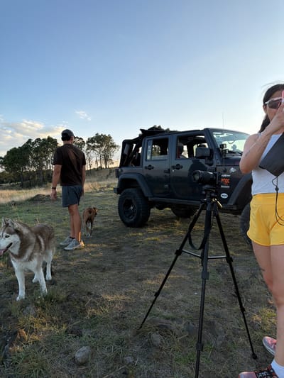 A photo taken outdoors shows two people, two dogs, a vehicle, and camera equipment. One person, wearing yellow shorts, a light-colored shirt, and pink sneakers, stands near a black Jeep with its top removed and a camera mounted on a tripod. The camera faces away from this person. The vehicle is on rough terrain with sparse grass. Another person wearing a dark shirt and gray shorts is standing with the dogs. One dog is a husky with white and brown fur; the second dog is tan in color. The sky is mostly clear, and trees are visible in the background. The location is in the city of Cholula, Mexico.