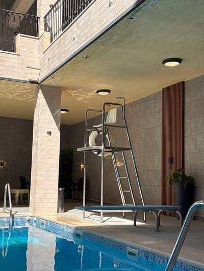 A low-angle, medium shot captures a swimming pool area at the JW Marriott Hotel in Ayn al Basha, Jordan. The scene is dominated by the bright blue, tiled edge of the pool in the foreground, with the water reflecting the overhead structure and sunlight. A metallic ladder with a life preserver attached rises from the pool area towards a raised seating platform. To the right of the ladder, a tall, dark pot holds a green plant, situated against a wall clad in dark, rectangular tiles. A wooden slatted panel is installed vertically on this wall. Overhead, a textured ceiling, illuminated by recessed lights, casts dappled shadows onto the scene. The background reveals a multi-story structure with a balcony supported by a light-colored, brick-patterned wall. This building features a black metal railing. The overall impression is of a well-maintained, modern outdoor space.