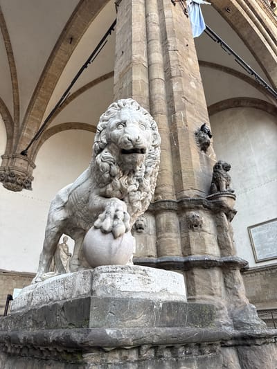 The image depicts the iconic Lion of Florence sculpture, also known as Marzocco, a sculpture of a lion with a king's crown, symbolizing Florence's power and sovereignty. This particular sculpture is a replica, as the original is housed in the Palazzo Vecchio museum. The replica stands on a stone pedestal in front of the Loggia dei Lanzi in Piazza della Signoria, Florence, Italy. The lion is depicted in a seated posture, with its right paw resting on a lily (Giglio), the symbol of Florence.
The sculpture is made of white marble, showing signs of weathering and age. It is positioned outdoors, with an ornate stone column and arched architectural features of the Loggia dei Lanzi in the background. A blue flag is visible hanging from the column. Pigeons are perched on ledges of the stone column, one with its wings slightly raised. In the background, through an archway and under the main sculpture, a small white marble statue of a standing figure can be seen. The ground surface around the pedestal appears to be paved with stone. The overall scene captures a moment of historical significance and artistic grandeur in the heart of Florence.