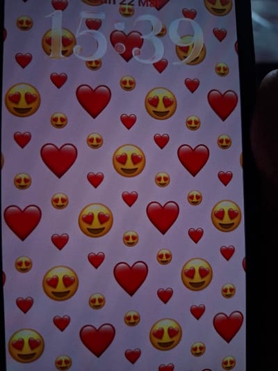 The image depicts a vertical close-up of a smartphone screen, situated within the city of Selyanin, Bulgaria. The specific point of origin within Selyanin is not discernible from the visual evidence.
The screen displays its lock interface, featuring a repeating pattern wallpaper composed of numerous red heart emojis and yellow smiling face with heart-eyes emojis, varying in size, set against a light pink background. Overlaid digitally on this pattern, in a large, light-colored font, is the time '15:39'. Above the time, the characters '22 M' are partially visible, consistent with a date display. A dark, out-of-focus object, consistent with a human thumb, grips the right edge of the device. The immediate surrounding environment of the phone is dark and indistinct.