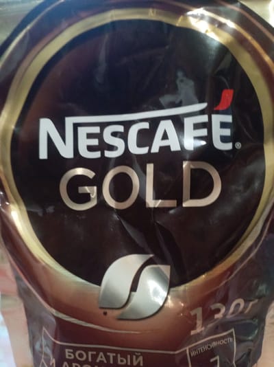The image shows a close-up of a Nescafe Gold coffee product packaging. The prominent elements are the "NESCAFE GOLD" logo in white and gold lettering, set against a dark, presumably brown, background with gold accents. There is a red curved logo near the "E" in "NESCAFE." Below the main logo, there is a silver stylized leaf-like emblem and text that appears to be in Russian. To the right of this emblem, the text "130г" is visible, indicating a weight. Further down, the word "ИНТЕНСИВНОСТЬ" is visible, meaning "intensity" in Russian. The packaging appears to be shiny and possibly creased. There is no identifiable context of a city, location, or environment beyond the product itself. The image is a product shot and does not depict a scene in Пралетарскі сельскі Савет, Belarus, or any other geographical location.