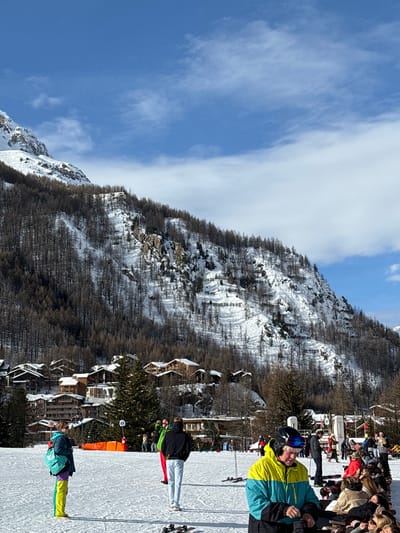 This media file captures a vibrant winter scene at a ski resort, specifically Val-d'Isère, France, under a clear blue sky with scattered white clouds. The image is taken outdoors during bright daylight, suggesting mid-morning to afternoon, with strong sunlight casting shadows on the snow-covered slopes.
In the foreground and midground, a ski slope is visible, populated by several individuals in ski gear. A person in a distinctive two-tone yellow and blue ski jacket, wearing a black helmet and goggles, stands prominently in the center-right, holding ski poles. Further down the slope, another person in a black jacket and white pants is visible, along with a third individual in a green/teal jacket and bright yellow pants. Their skis and poles are clearly seen.
The background features majestic snow-covered mountains, adorned with numerous bare trees. Nestled among the trees on the lower slopes are several rustic, chalet-style buildings, indicative of alpine architecture. To the right of the image, beyond the ski slope, a large group of people gathers. Some appear to be seated at outdoor tables, suggesting a restaurant or cafe area, while others are standing and socializing. The overall atmosphere is lively and relaxed, reflecting a typical day at a popular ski destination. There is no discernible text visible in the image.