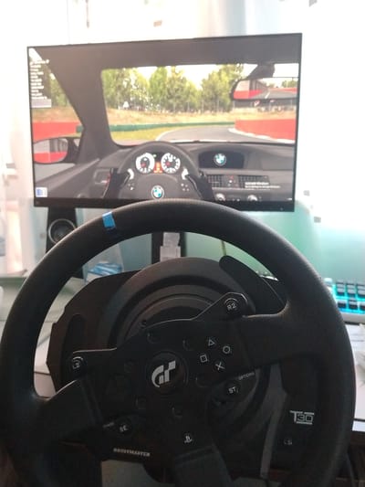 The image shows a gaming setup with a racing steering wheel and a monitor displaying a racing game. The steering wheel is black and features various buttons and a blue marker. The monitor displays a first-person view from inside a car on a race track, with trees and barriers visible. A speaker is located below the monitor. The background is blurred, but hints of a keyboard and other objects suggest a home or office environment. The image is not taken in Silistra, Bulgaria; it is a generic indoor scene. There are no people in the image.
