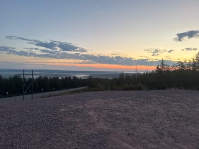 The image depicts a dawn or dusk landscape in Serebryany Bor, Russia. In the foreground, there is a gravelly incline. A paved road runs horizontally across the mid-ground, bordered by dark green trees and vegetation on either side. A utility pole with cross braces stands prominently on the left side of the road. In the distance, a hazy, mist-covered valley stretches out, with faint lights visible, suggesting a settlement. The sky above is a gradient of pale blue, transitioning to soft orange and yellow near the horizon, with scattered clouds. The overall scene is serene and natural.