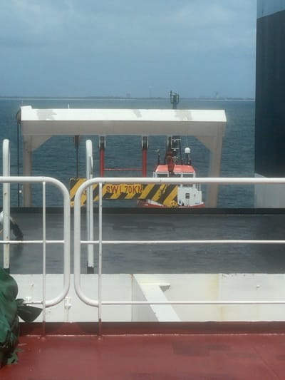 The scene captures a maritime perspective from the deck of a vessel, set off the coast of Orimedu, Nigeria. In the immediate foreground, a reddish-brown deck surface is visible, followed by white painted metal railings composed of horizontal bars and vertical supports, some forming U-shaped sections. Behind the railings, a dark grey deck surface extends. A white, gantry-like overhead structure spans the frame, from which a yellow and black striped lifting beam, marked "SWL 70KN" (Safe Working Load 70 Kilonewtons), is suspended by cables. Positioned beyond this equipment, a small white and red vessel, likely a tugboat or workboat, is afloat on calm, blue-green water. This vessel features a visible cabin, an antenna, and other deck-mounted structures. In the background, an expanse of water meets a distant, low-lying landmass on the horizon. Faint outlines of industrial structures and buildings, including a prominent vertical stack or tower, are discernible on the distant land. The sky is overcast with a light blue-grey hue. No individuals are present in the image.