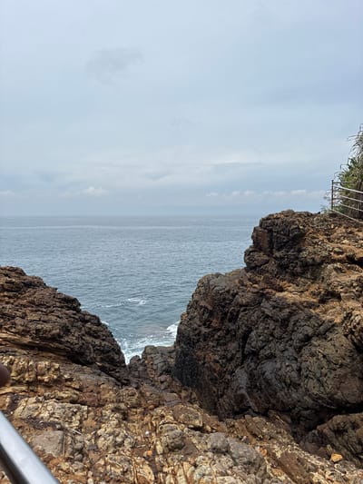 The image captures a view of the ocean from a rocky cliff in Dikwella, Sri Lanka. The sky is overcast, and the sea is calm with small waves. The cliff's rock formations are rugged and textured, with varying shades of brown and black. Vegetation and a metal railing are visible at the top left edge of the frame. The image is vertically oriented, offering a narrow glimpse of the sea through a gap in the rocks.