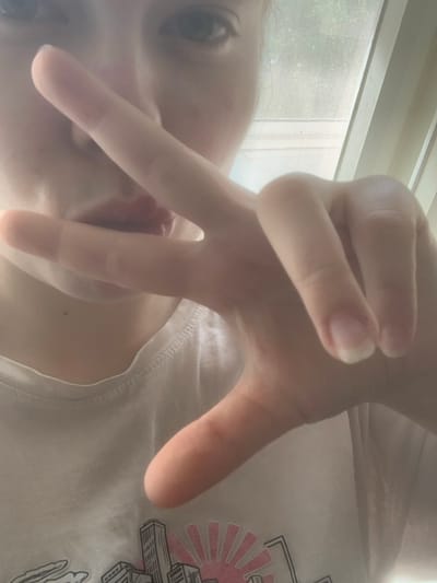 The image shows a person with fair skin and light-colored hair, possibly blonde, holding their hand up to the camera, fingers spread in a gesture that resembles a peace sign. They appear to be inside, as a window with a screen is visible in the background. The person is wearing a white T-shirt with a graphic print on the right side. The exact location in Novoe Devyatkino, Russia, is not discernible from the image.