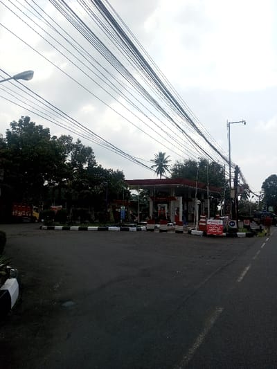 The image depicts an outdoor scene in the city of Niz Lakuk, India, featuring a gas station and its surrounding environment under an overcast sky. The foreground is dominated by a dark asphalt road with subtle lighter markings, leading towards the gas station. A small median with black and white curbing separates sections of the roadway.

The gas station, situated on the right-center, consists of a structure with a prominent red canopy supported by white pillars. Multiple fuel pumps are visible beneath the canopy, some displaying "Pertamina" branding. Several indistinct individuals are present near these pumps, appearing to be engaged in transactions or vehicle servicing.

To the left of the gas station, a cluster of dense green trees is visible, partially obscuring the background. A light-colored truck is parked among these trees on the far left. A tall, slender palm tree is situated directly behind the gas station canopy, towering over other foliage.

On the right side of the image, an individual wearing a dark shirt and light trousers walks away from the viewer on the edge of the road. A tall metal street lamp post with an attached security camera (CCTV) stands prominently on the right, next to various utility poles heavily strung with numerous overhead electrical or communication wires that traverse the upper half of the image. Another street lamp is visible on the far left, also accompanied by numerous overhead wires.

Multiple signs are present near the gas station entrance, including a red and white promotional sign and other directional or regulatory signs, one indicating a left turn and another appearing to be a "STOP" sign. The overall environment suggests a bustling, functional area.