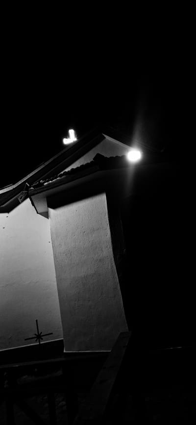 This is a low-light, black and white, vertical-format photograph of a building at night. The building appears to be a religious structure, possibly a church, given the presence of a cross on the roof and another cross-like symbol on the wall. A bright light fixture illuminates the roofline and casts shadows. The background is completely dark, suggesting the photo was taken in an outdoor environment with minimal ambient light. The overall scene is stark and possibly evokes a sense of solitude or solemnity. The image does not contain any people or depict any actions or interactions. The location in Kostenets, Bulgaria, is not visually evident from the image itself but is provided as context.