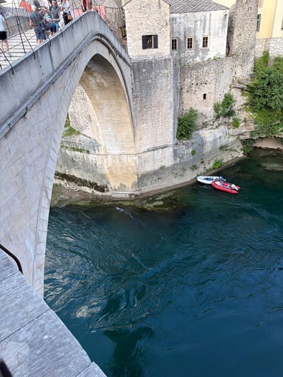 The image shows an elevated view of the Stari Most bridge in Mostar, Bosnia and Herzegovina, with the Neretva River flowing below. The stone bridge arch dominates the left side of the frame, with textured stone blocks and a metal railing visible on the walkway. Several people are walking on the bridge. On the right side, the river is a deep, dark blue, with two inflatable boats moored near the bank. One boat is red, and the other is white. The riverbank is rocky and overgrown with vegetation. The background features ancient stone buildings with arched windows and tiled roofs, characteristic of Mostar's historic architecture. The overall scene depicts a sunny day with clear water and a bustling atmosphere on the bridge.