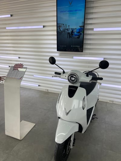 The image depicts the interior of a VinFast dealership or showroom in Kon Tum, Vietnam. A white VinFast electric scooter is prominently displayed in the central foreground, angled slightly left. It features a round headlight, black handlebars with rearview mirrors, and a distinct "V" emblem on its front fairing. The floor is light gray tile. The background wall is white with horizontal grooved panels, featuring embedded linear LED light strips and two visible electrical outlets. Mounted vertically on this wall is a large digital screen displaying "VINFAST MOBILE SERVICE" in English and Vietnamese, alongside images of individuals and VinFast scooters, with contact information and a website address. To the left, a white pedestal-style display stand holds a clear acrylic document holder with text, including "Bảo hành đến 8 năm" (Warranty up to 8 years). A person's hand is partially visible on the far left, near this stand.