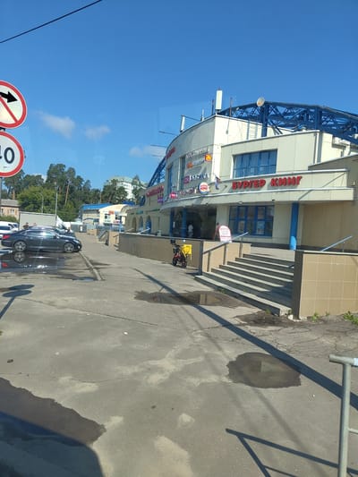 This is a photograph of a commercial building in Zhukovsky, Russia, on a clear day with a blue sky and some clouds. The building appears to be a shopping center or market, with various storefronts visible. One prominent sign reads "БУРГЕР КИНГ" (Burger King). Other signs include "ЗОМЯН" (presumably a store name) and "ОК ОПТИКА" (OK Optics). The building has a light-colored facade and a blue, modern-looking roof structure.
In the foreground, there is a paved area with several puddles on the ground, suggesting recent rain or cleaning. A dark-colored sedan is parked on the left side of the image. To the right of the building's entrance, there are a set of stairs leading up to the building. A red scooter with a yellow delivery box is parked near the bottom of the stairs. Two people are walking away from the camera on the sidewalk in front of the building.
In the upper left corner, two traffic signs are visible. One is a circular red and white sign with a black arrow pointing right, indicating a prohibition. Below it is a circular sign with the number "40" in black on a white background, likely indicating a speed limit. In the background, trees and other buildings are visible, including a taller, light-colored building in the distance. The overall scene depicts an outdoor urban environment.