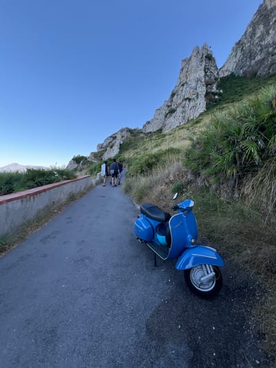 A blue Vespa scooter is parked on the side of a paved road in Sperone, Italy. The road curves uphill, flanked by a low brick wall and dense, dry vegetation. In the background, a group of four individuals walks up the road. Towering cliffs with sparse vegetation rise steeply on the right side of the image, under a clear blue sky. In the distance, on the left, a hazy mountain range is visible. The lighting suggests it is either early morning or late afternoon.