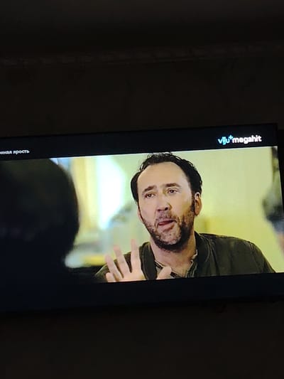 The media file shows a close-up of actor Nicolas Cage on a television screen. He appears to be in the middle of a scene, with his mouth open as if speaking, and a hand raised in a gesture. He has a beard and is wearing a dark jacket over a lighter-colored shirt. The background is blurred, with soft yellow and white lighting, suggesting an indoor setting. In the upper right corner of the screen, the text "viju+megahit" is visible, indicating the streaming service or platform. On the left side of the screen, there is some text in what appears to be Cyrillic script, partially obscured by a dark silhouette in the foreground. The overall impression is that of a paused moment from a film or television show. The context of Bokhtar, Tajikistan, does not appear to be directly relevant to the visual content of the screen itself.
