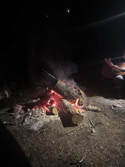 The image depicts a campfire scene at night, presumably in a rural setting. A person is visible in the top left, holding a guitar. The focus is on the campfire, which consists of logs burning brightly, with visible flames and smoke. The ground around the fire is covered in dirt, dry leaves, and twigs. The background is dark, with a faint light source visible in the distance on the left. No specific details identify the location as Юзуфоўскі сельскі Савет, Belarus; the scene is a generic outdoor campfire setting.