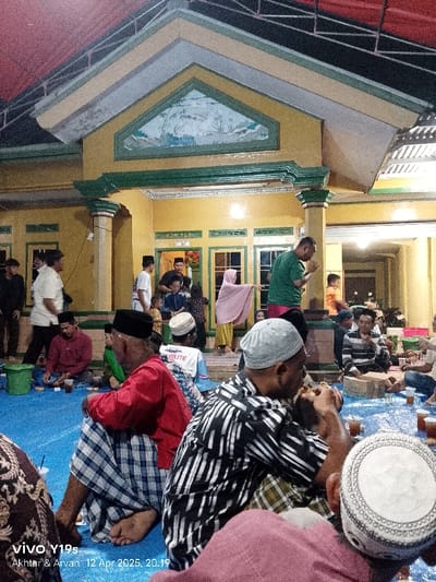 The image shows a gathering of people at a home in Anjatan, Indonesia (latitude: -6.419, longitude: 107.978). The home is a two-story structure with a yellow facade and a green trim. The front porch is covered by a red and black awning.  There are 15 people visible in the image. Most of them are men. They are all wearing traditional clothing.  They are seated on a blue tarp, drinking and eating. There are several empty glasses and food containers on the tarp. Two men are standing and talking to each other. There is a woman in a white headscarf standing on the porch. The man closest to the camera is wearing a black and white striped shirt. He is eating something with his right hand. The man next to him is wearing a red shirt and plaid pants. He is holding a glass in his left hand. The other people in the image are mostly obscured by the men in the foreground.  There is a white, plastic bucket in the foreground. There is a sign on the wall of the home that reads "Viva Y19s". There is another sign on the tarp that says "Akhtar & Arvan 12 Apr 2025, 20:19". The image is taken at night, but the lights are on inside the home and on the porch. The air is clear.