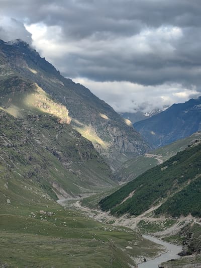 This image depicts a mountain valley scene in Gramphu, India. The valley is carved by a meandering river that flows through the foreground and mid-ground. Steep, rocky mountain slopes flank the river on both sides. The left slope is partially illuminated by sunlight breaking through dramatic, dark clouds, highlighting patches of greenery and rocky outcrops. The right slope is densely covered with dark green vegetation, with a winding road visible traversing its side. In the distance, more mountain ranges are visible, some with snow-capped peaks shrouded in mist. The sky is filled with heavy, grey clouds, with shafts of light piercing through in places. The overall impression is one of a vast, rugged, and somewhat dramatic natural landscape under a dynamic sky.
