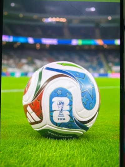 This is a close-up, vertically oriented photograph of a soccer ball on a vibrant green grass field. The ball features a distinctive design with white panels adorned with blue stars and a prominent FIFA logo. Red and green accents are also visible on the ball's panels. The background is blurred, showing stadium seating with colorful lights and a hint of the crowd. The lighting suggests an indoor or evening event, as indicated by the artificial lights in the background. No people are clearly discernible in the image. The text "FIFA" and "TRION" are visible on the ball. The setting appears to be a modern sports stadium.