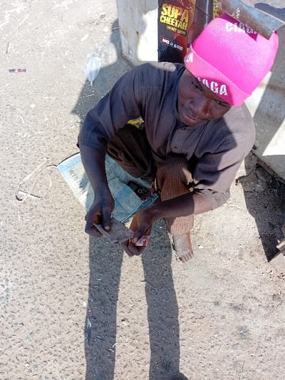 In Sharada, Nigeria, an adult male with dark skin is observed squatting outdoors on a rough concrete and dirt surface under bright sunlight. He wears a dark brown long-sleeved tunic, dark brown striped trousers, and a pink knitted beanie with white text. He is barefoot. His hands are engaged in handling several folded currency notes, likely Nigerian Naira. A light blue and white patterned sack or fabric is positioned beneath his left side. Behind him, a partially visible commercial poster advertising 'SUPA CHEETAH ENERGY DRINK' is affixed to a light-colored vertical structure. Elongated shadows from the individual and unseen objects are cast on the ground, which also contains minor debris including a clear plastic bag.
