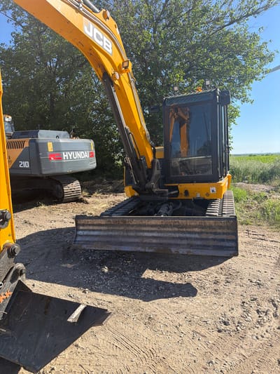 A partially obscured Hyundai 210 excavator and a JCB excavator are parked on a dirt road adjacent to a field, with a tree partially obscuring both pieces of machinery. The location is in the vicinity of Selanovtsi, Bulgaria.