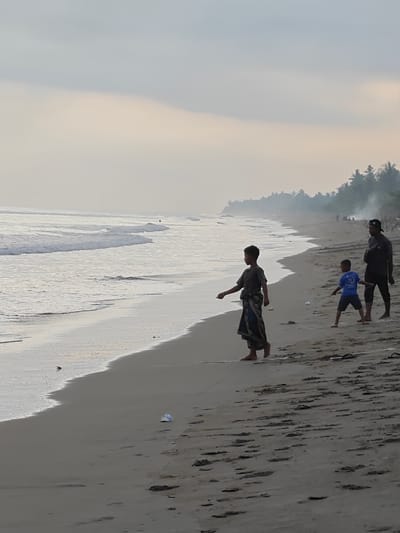 A group of three people, two children and one adult male, stand on the shore of a beach in Curehtunong, Indonesia. The children are in the foreground, with one child wearing a sarong and the other wearing a blue t-shirt. The adult male stands behind them, wearing a dark shirt and a baseball cap. The ocean is to the left of the frame, with waves gently rolling onto the shore. In the background, a line of palm trees and smoke suggest a village or settlement along the coastline. The sky is overcast, with soft light filtering through the clouds. The overall scene depicts a serene moment on a tropical beach.