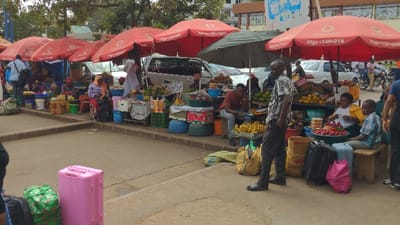 A bustling outdoor market scene unfolds. Several vendors sit under red umbrellas, selling fruits and vegetables. Cars are parked behind the stalls. A man in a patterned shirt stands near bananas and suitcases. Additional luggage rests on the ground near a pink suitcase. Other individuals are visible, some seated at the market stalls, possibly as customers.