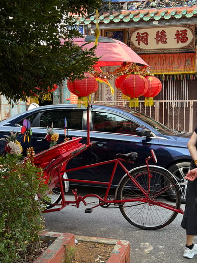 A parked blue sedan, with a red bicycle strapped to it, sits in front of a building adorned with Chinese lanterns and characters, located in George Town, Malaysia. Three red paper lanterns hang from the vehicle's front end, decorated with tinsel. Flowers and pinwheels adorn the car. A person wearing black pants and white shoes stands near the rear wheel of the car, facing away from the camera. Bushes partially obscure the car and bicycle. A planter with a red curb is visible on the right side of the image, bordering the sidewalk.