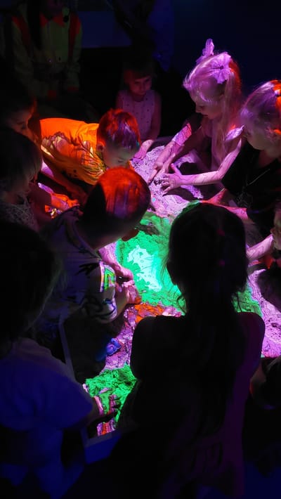 This image depicts a group of children gathered around a brightly lit, sand-filled table or interactive exhibit, likely in Komsomolsk-on-Amur, Russia. The exhibit glows with vibrant green and purple light, illuminating the children's faces and hands as they interact with the sand. The children, varying in age, are leaning in, touching, and exploring the textured surface, their expressions suggesting curiosity and engagement. The surrounding environment is dark, accentuating the illuminated exhibit and the children's involvement. The overall scene suggests an educational or recreational activity designed to stimulate sensory exploration and imaginative play.