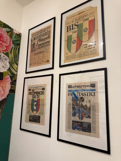 The image shows four framed newspapers mounted on a white wall. The newspapers are in Italian and appear to be sports-related, with headlines and images about the Napoli football club. The newspapers are arranged in a grid of two columns and two rows.
The top left newspaper features a headline that translates to "Napoli champion" and an image of a jubilant person. The top right newspaper has a large "BIS" (meaning "again" or "second") with the years "1987" and "1990," and a graphic of two shields with the Napoli crest. The headline reads "IL MATTINO" and "Città di mare con abitanti con scudetti," which translates to "City of the sea with inhabitants with league titles."
The bottom left newspaper has the headline "RICOMINCIO DA TE," meaning "I start again from you," and a large number "3" within a shield. The bottom right newspaper is a special edition of "IL MATTINO" with the headline "NAPOLI CAMPIONE D'ITALIA" (Napoli Champion of Italy) and "FANTASTICI" (Fantastic). It shows images of the Napoli team celebrating.
A portion of a large artwork with floral motifs is visible on the far left of the image. The environment is indoors, likely a home or gallery, with a plain white wall. There is no information in the image that places the scene specifically in the city of Naples, Italy, but the content of the newspapers strongly suggests a connection to the city and its football team.