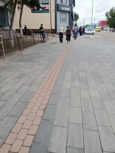 A young man in a dark t-shirt and jeans is leaning against a metal fence, looking at his phone. He is standing on a sidewalk made of grey and reddish-brown paving stones. The sidewalk is adjacent to a building with a beige facade and trees. In the background, there are other people walking on the sidewalk and street, as well as cars and a yellow bus. The scene appears to be a commercial or residential area in the city of Kostanay, Kazakhstan. The sky is overcast.