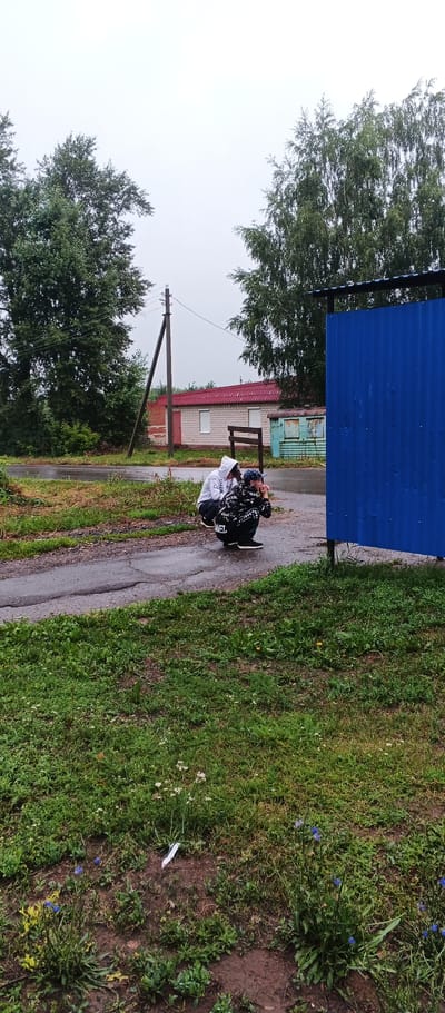 Two young males are squatting on a wet, paved surface in front of a vibrant blue corrugated metal structure. The male on the left is wearing a white hooded sweatshirt, and the male on the right is wearing a black hooded sweatshirt with white lettering and a baseball cap. They appear to be looking at something to the right of the frame. The environment is outdoors with lush green grass and various trees visible in the background. A telephone pole stands to the left, and a building with a red-tiled roof is situated behind the men. The sky is overcast. The location is identified as the city of Podgornoye, Russia.