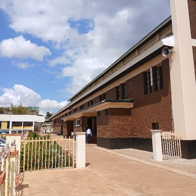 The image depicts an outdoor scene featuring buildings, sky, and ground, captured on a bright day. The primary subject is a multi-story building with reddish-brown brick walls and light beige or off-white trim, extending horizontally across the frame from left to right. This building features numerous dark-paned windows, some appearing to have internal blinds or coverings. A continuous, light-colored gutter or overhang system runs along the roofline on the left side of the building. A dark, dome-shaped security camera is mounted under this overhang towards the left.
In the foreground and to the right, there is a wide expanse of light brown or reddish-brown paved ground. Another section of a building, similar in construction to the main one, is visible on the right, featuring a small, light-colored railing or balcony. One person is standing near this right-hand building, wearing a dark top and light pants, appearing to look down or away from the camera. Further to the right and in the lower foreground, a prominent yellow metal gate with vertical bars and some decorative elements is visible, behind which green foliage and bushes can be seen, indicating landscaped areas.
A significant portion of the upper part of the image is filled with a bright blue sky dotted with fluffy white cumulus clouds, suggesting clear and sunny weather. The overall lighting indicates it is daytime, likely mid-morning or afternoon. No discernible text is visible in the image. The scene conveys a calm, everyday outdoor environment.